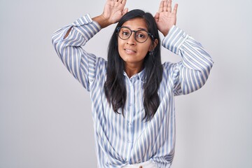 Young hispanic woman wearing glasses doing bunny ears gesture with hands palms looking cynical and...
