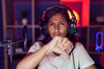 Young hispanic woman playing video games wearing headphones with angry face, negative sign showing dislike with thumbs down, rejection concept
