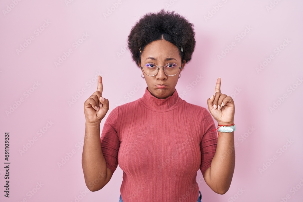 Sticker beautiful african woman with curly hair standing over pink background pointing up looking sad and up