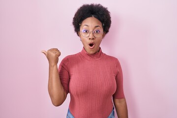Beautiful african woman with curly hair standing over pink background surprised pointing with hand finger to the side, open mouth amazed expression.