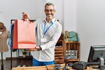 Middle age grey-haired man shop assistant holding shopping bag at clothing store - obrazy, fototapety, plakaty