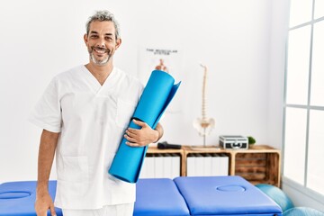 Middle age grey-haired man physiotherapist smiling confident holding yoga mat at rehab clinic