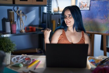 Fototapeta na wymiar Young modern girl with blue hair sitting at art studio with laptop at night smiling with happy face looking and pointing to the side with thumb up.