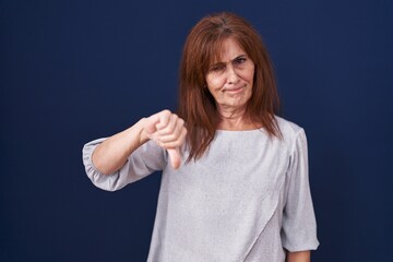 Middle age woman standing over blue background looking unhappy and angry showing rejection and...