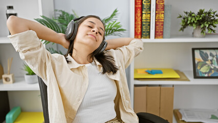 Young, beautiful, hispanic woman employee, joyful and confident, tuning into music indoors at the office. relaxed at her executive desk, hands on head, she values the sound of success!