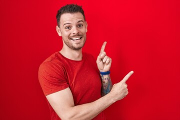 Young hispanic man standing over red background smiling and looking at the camera pointing with two...