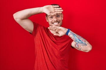 Young hispanic man standing over red background smiling cheerful playing peek a boo with hands...