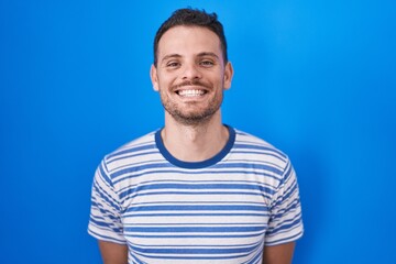 Young hispanic man standing over blue background with a happy and cool smile on face. lucky person.