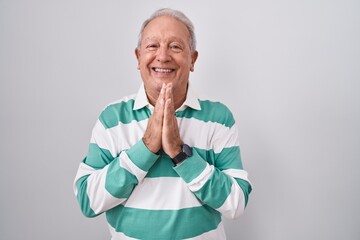 Senior man with grey hair standing over white background praying with hands together asking for...