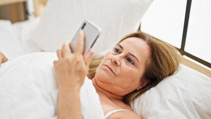 Middle age hispanic woman looking smartphone lying on bed at bedroom