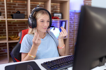 Young caucasian woman playing video games wearing headphones doing money gesture with hands, asking...