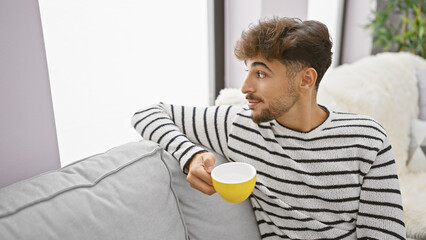 Relaxing morning, handsome young arabian man with a beard, comfortably resting on his sofa at home, enjoying his cup of espresso, indulging in the simple pleasure of a quiet, coffee drink