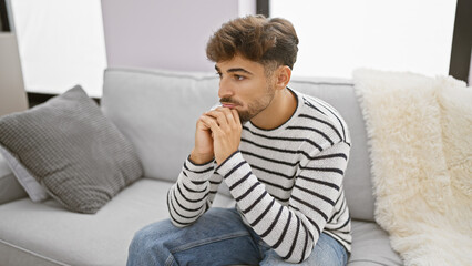Worried young arab man with a beard, sitting on his living room sofa at home, seriously expressing...