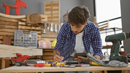 Diligent young arabian man jotting down ideas on touchpad in carpenter's workshop, sculpting his...
