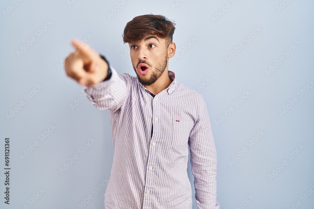 Wall mural arab man with beard standing over blue background pointing with finger surprised ahead, open mouth a