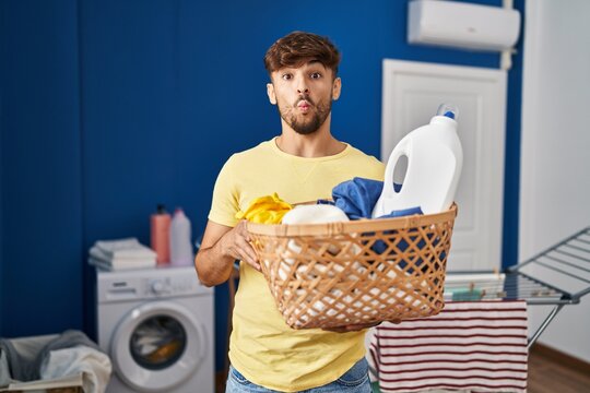 Arab man with beard holding laundry basket and detergent bottle making fish face with mouth and squinting eyes, crazy and comical.