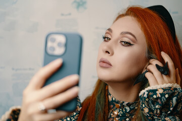 Female blogger shoots herself for a blog about fashion and makeup. Red-haired girl takes a selfie on the front camera of a modern smartphone. Adorable woman with a ethnic slavic crown on her head.