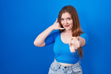 Redhead woman standing over blue background smiling doing talking on the telephone gesture and pointing to you. call me.