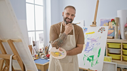 Cheery, confident young man, artist with a beaming smile, joyfully painting and holding his palette...
