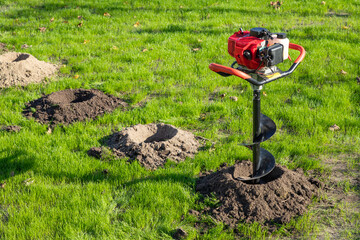 Tool hand-held soil hole drilling machine or portable manual earth auger for prepare the soil for planting shrubs or trees in newly drilled hole on a green lawn.