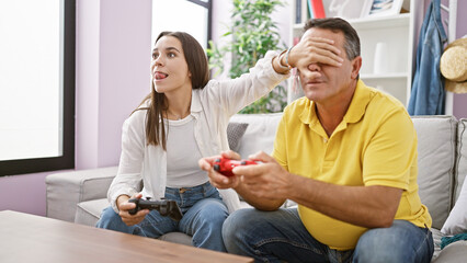 Fototapeta na wymiar Hispanic father and daughter enjoy playing video game on the cozy home sofa, covering eyes and hands in laugh-filled skulduggery