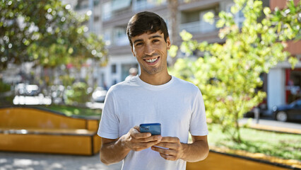 Cheerful young hispanic man joyfully using mobile technology, texting away in the heart of the city park. handsome guy smiling, lost in his digital world among nature's green outdoors.