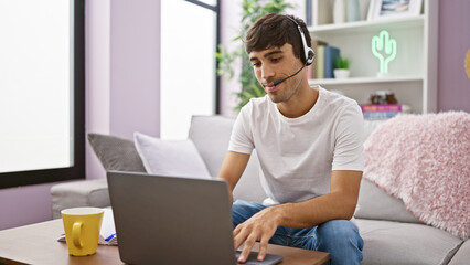 Young hispanic man call center agent using laptop and headphones sitting on sofa at home