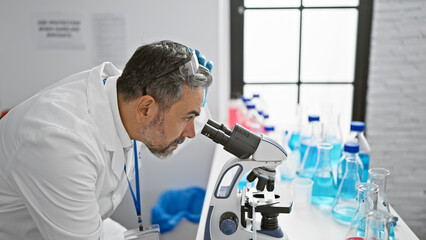 Naklejka na ściany i meble Attractive grey-haired young hispanic man, a dedicated scientist, working intently on a medical experiment in the lab, safety gloves on, peering through a microscope, deep in concentration.