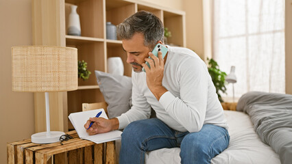 Handsome young hispanic man with grey-hair, engrossed in a serious phone talk, sitting on bed,...