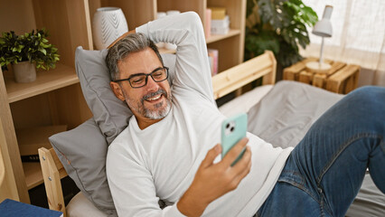 Beaming grey-haired young hispanic man lying on a cozy bed, engrossed in using his smartphone in a comfortable room, exemplifying the relaxed, confident lifestyle.