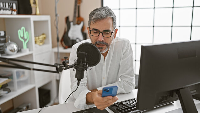 Grey-haired, young hispanic man, a radiant radio news reporter, passionately presenting stories and events, reading off smartphone screen at the bustling radio studio.