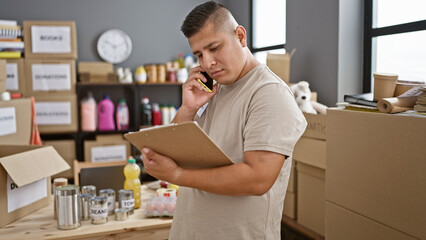 Fototapeta premium Confident hispanic young man standing at charity center, volunteering his service, reading donation documents while smiling and having a warm conversation on his smartphone