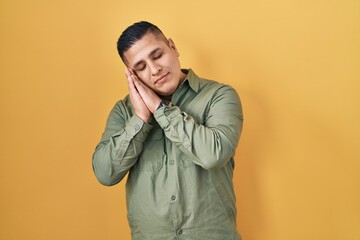 Hispanic young man standing over yellow background sleeping tired dreaming and posing with hands together while smiling with closed eyes.