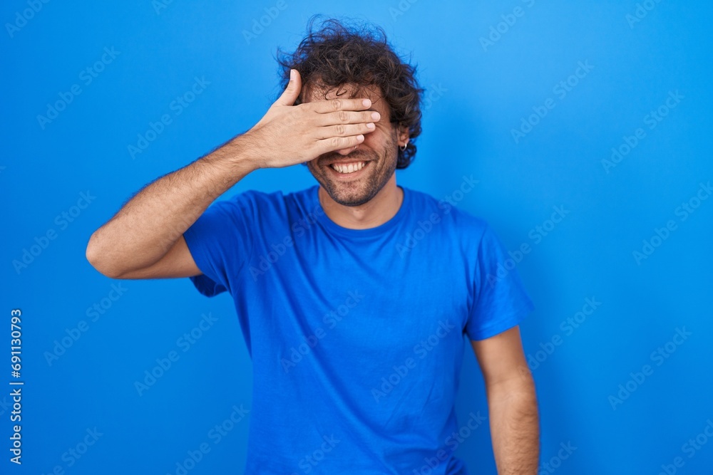 Sticker hispanic young man standing over blue background smiling and laughing with hand on face covering eye