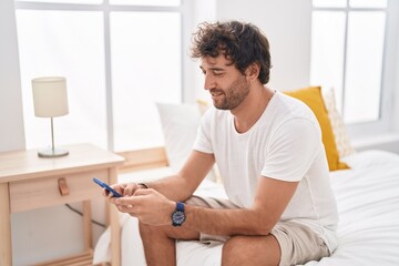 Young hispanic man using smartphone sitting on bed at bedroom
