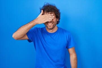 Hispanic young man standing over blue background smiling and laughing with hand on face covering eyes for surprise. blind concept.