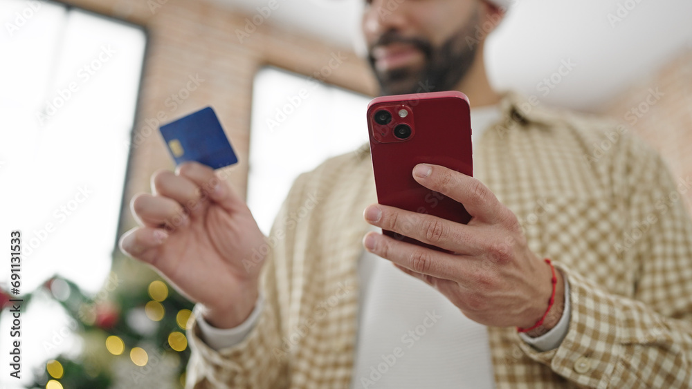 Sticker young hispanic man shopping with smartphone and credit card celebrating christmas at home