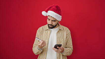 Young hispanic man using smartphone and credit card wearing christmas hat over isolated red background