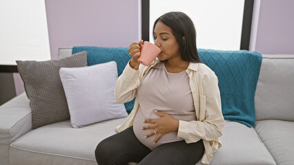 Obraz na płótnie Canvas Home comfort, young pregnant woman enjoying a relaxing morning at home, drinking coffee and touching her belly on a comfortable sofa