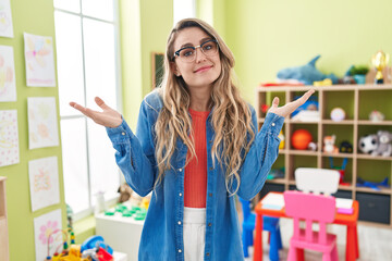 Young caucasian woman working as teacher at kindergarten clueless and confused expression with arms...