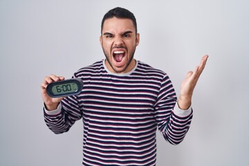 Fototapeta premium Young hispanic man wearing pyjama holding alarm clock celebrating victory with happy smile and winner expression with raised hands