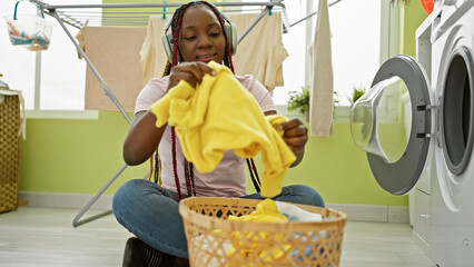 Joyous african american woman listening to soulful music, washing clothes with a gleaming smile in...