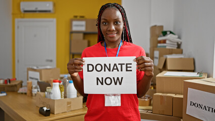 Beautiful braided african-american woman confidently smiling as she volunteers. holding up a donate now message at a charity center, showing unity and altruism through her service.