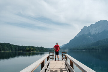 View from the shore of Lake Eibsee in Germany, with a hiker in a red shirt standing on a footbridge...
