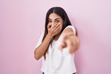 Young arab woman standing over pink background laughing at you, pointing finger to the camera with hand over mouth, shame expression - obrazy, fototapety, plakaty