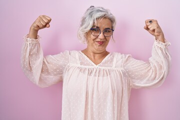 Middle age woman with grey hair standing over pink background showing arms muscles smiling proud....