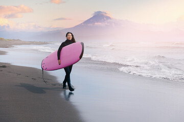 Extreme surfer happy young woman in wetsuit with surfboard go to winter surfing in Atlantic ocean Kamchatka Russia on background volcano - obrazy, fototapety, plakaty