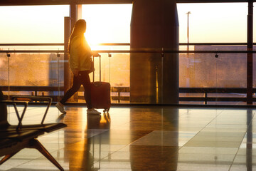 An airport passenger with a suitcase walks against the backdrop of the morning sun. View from the...