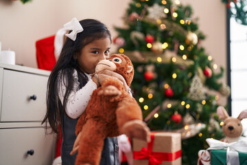 Adorable hispanic girl hugging and kissing monkey doll standing by christmas tree at home