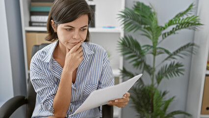 Pensive young hispanic woman immersed in paperwork, doubting amidst a sea of documents in her...
